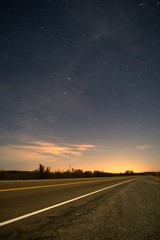 Asphalt road vanishes on the horizon at night under a deep, blue starry sky.