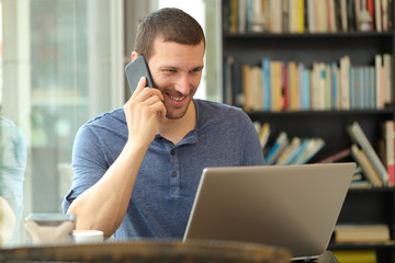 Happy man talks on phone checking laptop content in a bar