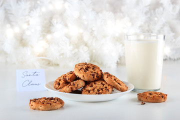 Handmade chocolate chip cookies and a glass of fresh milk are a gift for Santa Claus on Christmas day. The lights are on decorating the white Christmas tree. Christmas holidays concept