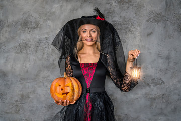 Young blonde woman in costume standing over grey background with pumpkin and lighter