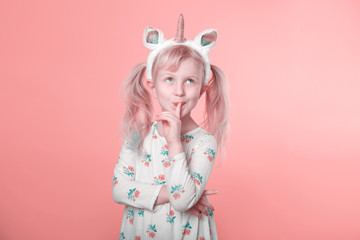 Cute adorable Caucasian blonde girl in white dress wearing unicorn headband horn and ears thinking dreaming. Funny kid child expressing emotion standing in studio on pink coral background.