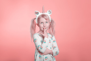 Cute adorable Caucasian blonde girl in white dress wearing unicorn headband horn and ears thinking dreaming. Funny kid child expressing emotion standing in studio on pink coral background.