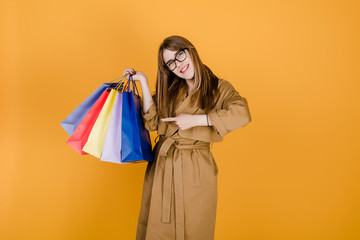 hipster young european woman in glasses and coat with colorful shopping bags isolated over yellow