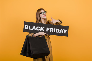 smiling woman in glasses with black friday sign and paper shopping bags isolated over yellow
