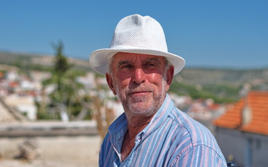 Waist up portrait of a senior man with a hat with cityscape of Primosten in Croatia in the background