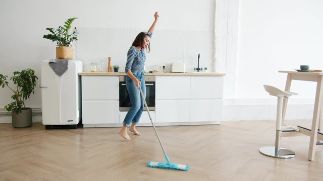 Happy young woman cute housewife is washing floor with mop and dancing at home in kitchen enjoying housework. People, modern lifestyle and youth concept.