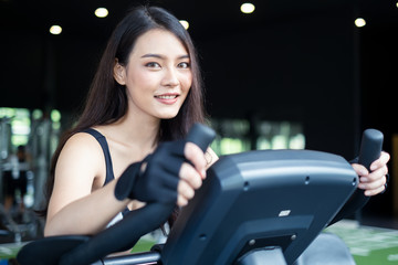 Fototapeta na wymiar Crop shot of Asian beautiful sporty young woman workouts in Gym or fitness club. The girl cycling on elliptical training machine and looking at camera with smile. Exercise for wellness and good health