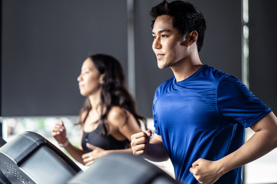 Young Fit Handsome Asian Man And Beautiful Asian Woman Running On Treadmill Or Running Machine In Modern Fitness Gym. Seen From Side View While They Focusing On Running. Workout In Gym And Fitness.