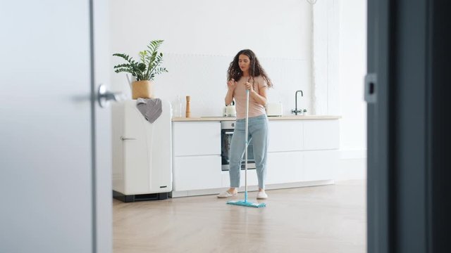 Joyful girl with long curly hair is dancing with plastic mop in kitchen singing enjoying music during clean-up at home. Creative youth and lifestyle concept.