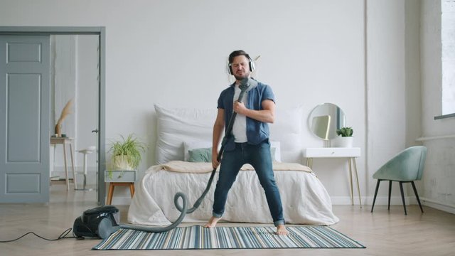 Happy young man in headphones is singing in vacuum cleaner dancing pretending to play the guitar cleaning home alone. Lifestyle and housework concept.