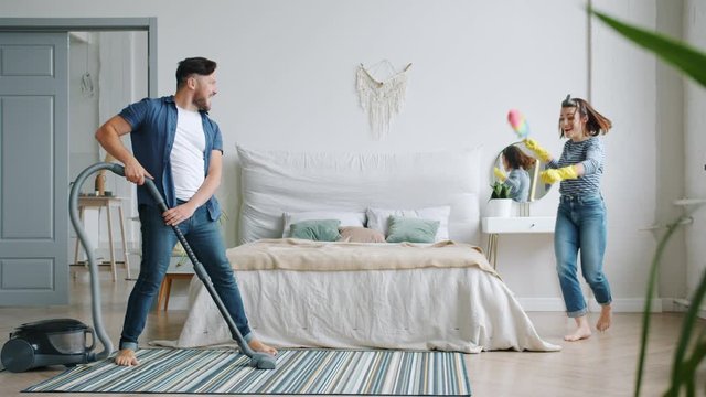 Housewife pretty woman is dusting furniture then singing in duster and dancing with husband and vacuum cleaner. Music, lifestyle and happiness concept.