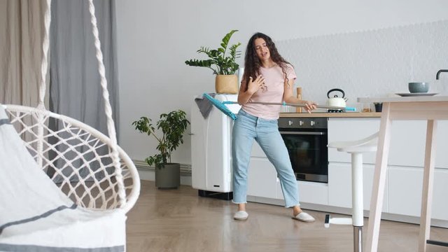 Slow motion of silly student dancing with mop in kitchen enjoying music relaxing during clean-up in studio apartment. Youth and modern lifestyle concept.