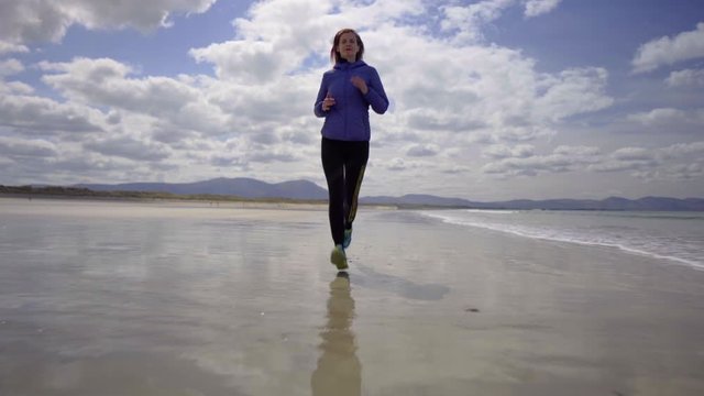 Low dolly shot of a girl running, jogging on the shore of a sandy beach with Atlantic ocean waves on a wonderful sunny day in Ireland in 4K