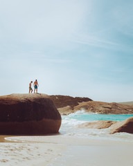 Two adventurous men in board shorts, on top of a huge boulder overlooking the beautiful blue waves of the ocean, at Little Beach, Albany, Western Australia. 