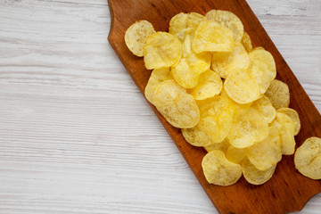 Yellow potato chips with salt on a wooden board on a white wooden surface, top view. Flat lay, overhead, from above. Space for text.