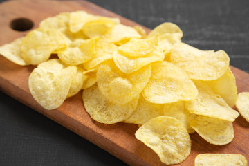 Delicious potato chips with salt on a rustic wooden board on a black surface, side view. Closeup.
