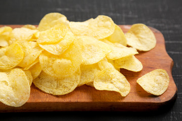 Delicious potato chips with salt on a rustic wooden board on a black surface, side view. Close-up.