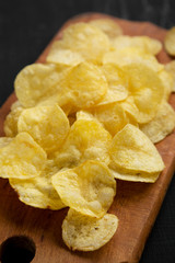 Crispy potato chips with salt on a rustic wooden board on a black surface, side view. Closeup.