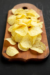 Crispy potato chips with salt on a rustic wooden board on a black surface, side view.