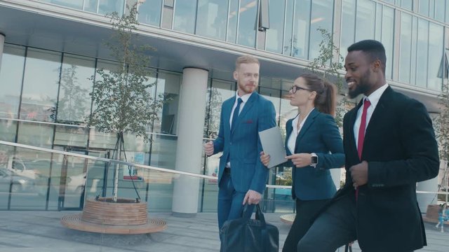 Male and female business people walk and discuss business. Group of colleagues going along modern glass business center. Three confident persons in formal suits