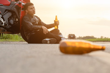 Motorcyclist sitting on the road beside his motorcycle and drinking an alcohol or beer