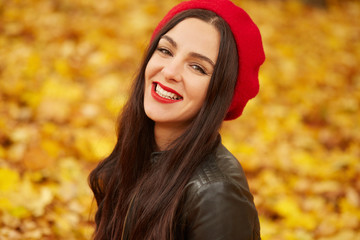 Fototapeta premium Close up portrait of autumn woman in autumn park, spending sunny days in fall forest, girl wearing red beret and black leather jacket, looking smiling directly at camera. People concept.