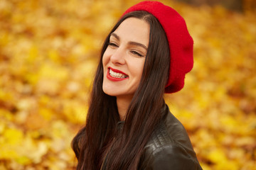 Happy beautiful woman, fashion model having fun in fall park, looking smiling away. Outdoors young pretty woman in autumn park, dresses red beret and leather jacket, female being in good mood.