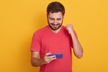 Photo of delighted man dresses red casual t shirt playing video game on cellphone and clenching fist isolated over yellow studio background, bearded guy posing in studio. People and technology.
