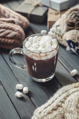 Hot cocoa with marshmallows in a glass cup surrounded by Christmas gifts and winter clothes, Christmas atmosphere and coziness