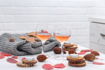 Small candles, two glasses with rose wine, cones, dry red leaves, a gray scarf knitted on a white wooden table. Hello, Autumn. Cozy autumn background.