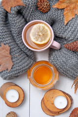 A cup of tea with lemon and honey, a gray woolen scarf, cones, dry leaves, candles on wooden coasters on a white, wooden table. Autumn comfort.