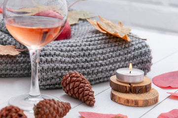 Fototapeta na wymiar Small burning candles, two glasses with rose wine, cones, dry red leaves, a gray scarf knitted on a white wooden table. Hello, Autumn. Cozy autumn background.
