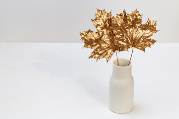 Simple white ceramic vase with leaves of marple tree. Gold colored leaves on white table, autumn time. Modern home decoration bouquet . Minimal still life.