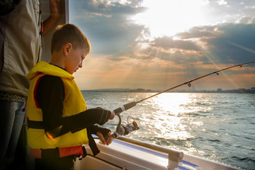 Little boy fishing on a fishing rod from a boat at sunset