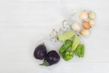 Green bell peppers, round onions and eggplants in eco natural bags sewing from mesh cloth on light background with copy space. Zero waste food shopping concept. View from above.