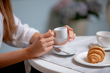 Side view of female hands holding cup of coffee. Woman having coffee brake in French cafe. Women drinking coffee in French styled restaurant. Croissants and coffee on white table.