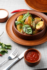 Fried potato in brown clay bowl close-up. Front view of plate with fried potato and basil with sauces and fork with knife. Focus on fried potato. Grey background. vertical photo