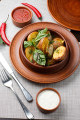 Fried potato in brown clay bowl close-up. Front view of plate with fried potato and basil with sauces and fork with knife. Focus on fried potato. Grey background. vertical photo