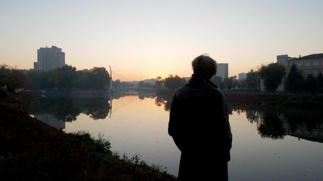 Silhouette of man in black coat standing alone near river and city with sunrise sky