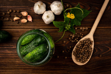 Fresh cucumbers in glass jar, garlic and black pepper on dark wooden background.