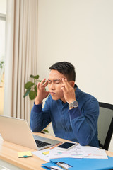 Feeling exhausted. Frustrated young beard man massaging his head and keeping eyes closed while sitting at his working place in office