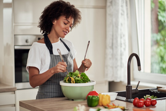 Attractive mixed race woman in apron and with curly hair mixing vegetables in bowl while standing in kitchen at home.
