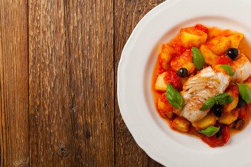 Traditionally prepared cod in Spanish. Served in tomato sauce with boiled potatoes, olivki and basil. Top view.