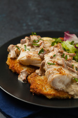 Fried cod, served on potato pancakes with mushrooms sauce and salads. Portion on a black plate. Dark background.