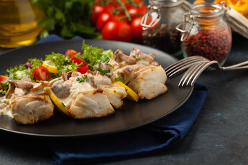 Fried cod served with mushroom sauce and salads. Portion on a black plate. Dark background.