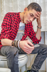 Young man is sitting drunk on a toilette ready to swallow drug pills