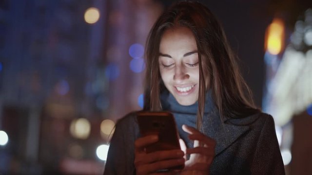 Closeup of attractive smiling young woman using smartphone walking outdoors night autumn city, slowmotion fullHD stock footage, handheld camera shot. female model messaging, texting, swiping screen on