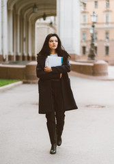 Fototapeta premium Beautiful serious smart girl brunette student holding notebooks and textbooks, goes walking at University on street of St. Petersburg. A charming woman with long dark hair is studying at course