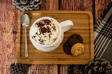 Hot chocolate with whipped cream sprinkled with cocoa on a white mug with a spoon and cookies on a side standing on a bamboo plate with pinecone and wooden background