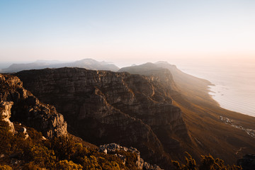 The view from on top of Table Mountain, Cape Town, South Africa. Beautiful mountainous landscape with the sea and iconic bays in the distance. The golden sun is setting over the ocean horizon. 
